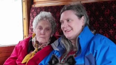 Miriam Souter has short grey hair and is wearing a pink coat and yellow scarf. Sat next to her is her daughter Phazar, who has platted brown hair and is wearing a blue coat. On Phazar's lap is a medium sized black dog.