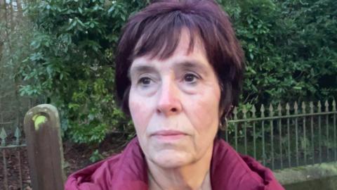 A woman wearing a purple coat stands with a solemn look on her face in front of metal railings and a green leafy bush. 