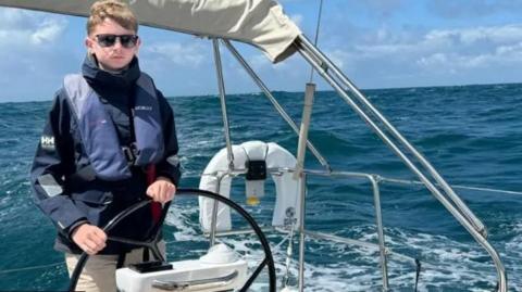 A teenager dressed in a blue sailing jacket and blue lifejacket is at he helm of a yacht. He has sun glasses on and there are some big waves behind him.