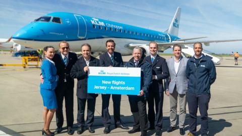 A picture of a number of people stood in a line. A number of people are wearing suits. There is also a sign held up by three people which reads 'New flights Jersey - Amsterdam'. There is a large blue and white KLM plane that they are stood in front of.