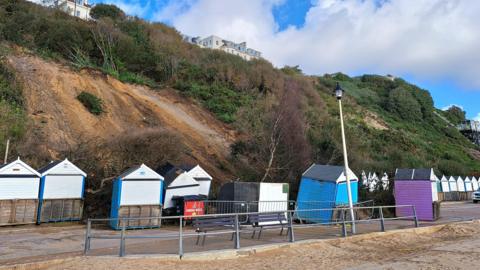 View of the cliff slide that has impacted the beach huts. At least four of them have been displaced. Buildings can be seen on top of the cliff. It's a cloudy day.