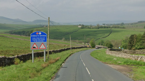 A dales view with a large blue sign that reads 'Dibble's Bridge Long, steep descent. Cyclists test brakes & drive carefully'. 