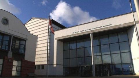 Douglas Courthouse, a large pale-coloured court building with a glass front. A flagpole flying a Manx flag stands in front of it on the left hand side.