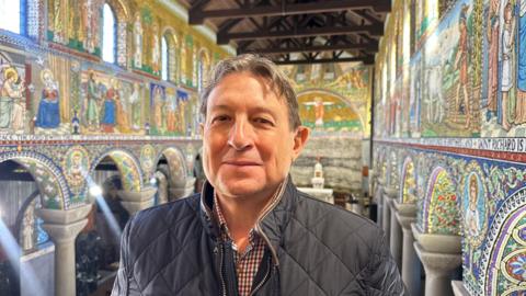 Vella is pictured inside the church with mosaic walls behind him. He wears a dark quilted jacket.