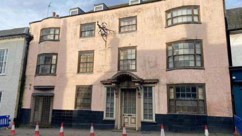 An old cream building with multiple windows and doors. There is a row of cones in front of the building. 