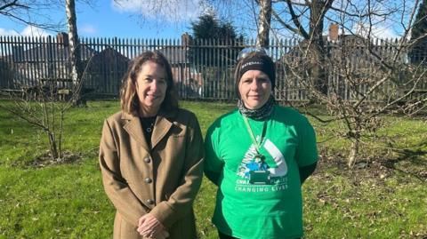 Joe Chadwick-Bell wearing a brown coat next to Melissa Underwood, in a green t-shirt standing in a grassy area in front of a silver metal fence