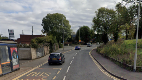 A road with a bus stop on the left hand side, with three cars driving on it. There are trees on the left and right hand side of the road