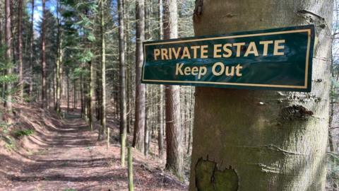 A green and yellow sign reading 'Private Estate Keep Out' is nailed to a tree trunk, with woodland trees and path out of focus behind.