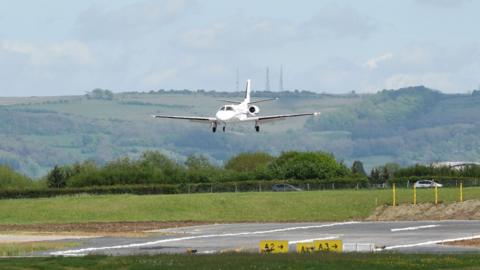 A small white plan landing at Gloucestershire Airport. There is a green hill behind covered in trees and fields and the sky is blue. 