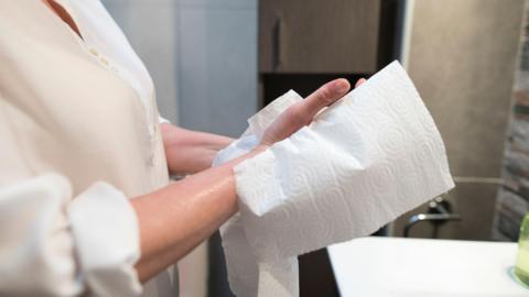 A close up image of a woman in a white shirt wiping her hands on a paper towel in a bathroom. 