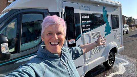 A woman with short, pink hair smiling with her arm out stretched in front of a campervan. On the side of the campervan is has a picture of Britain and says "Tracey's trek".