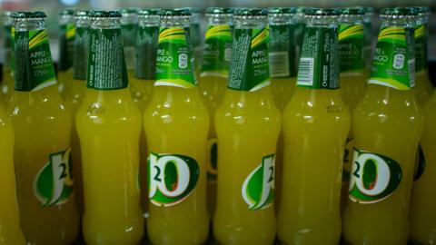 Lots of bottles of apple and mango flavoured J2O on a shelf. The drink is a faint orange in colour and the branding is white and green.