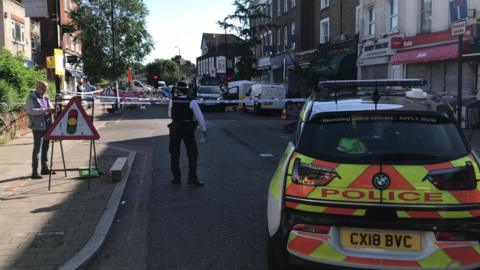 Police officers on Stroud Green Road where a blue and white police cordon blocks off the street. A police car is parked on the right hand side and on the left there is a traffic light road sign and a male passer by possibly taking a photo.