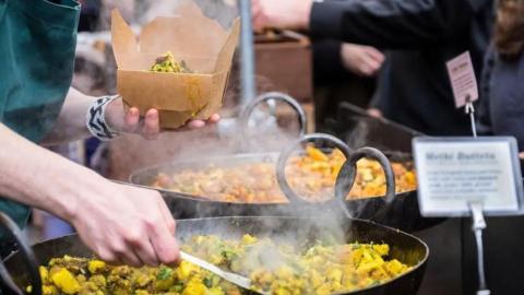 A person serving some street food from a large pan, putting it into a cardboard box.