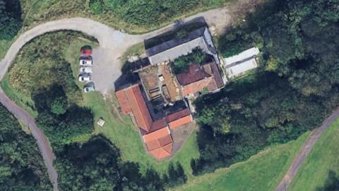 An aerial view of a farmhouse, surrounded by fields and cars on a driveway. 