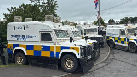 A number of Police Service of Northern Ireland landrovers are seen parked up beside a bridge in the village of Scarva ahead of the arrival of a Palestine solidarity parade. A Union Jack flag can also be partially seen at the top of the picture attached to a lamp post.
