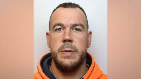 A custody image of Daniel Smith. He is staring directly into the camera and has short brown hair and a short beard and moustache. He is wearing an orange hooded top.