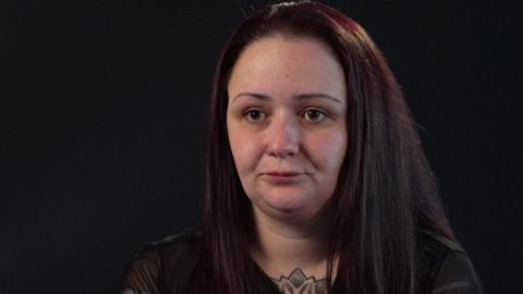 Hannah Mellor sat against a black backdrop. She has dark hair and is wearing a black top with a tattoo seen below her neck. 