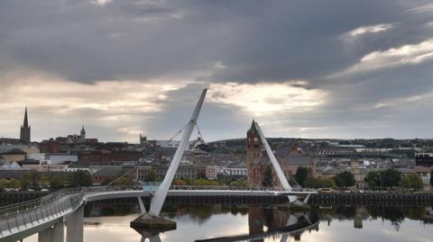Shows Derry's peace bridge beneath grey skies with the Guildhall, St Columb's Cathedral and other buildings in the background.