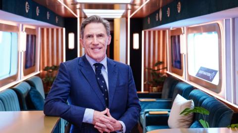 Rob Brydon standing in a blue suit in a carriage with blue seats behind him.