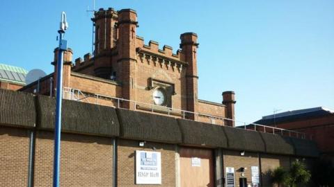 The main gate of Hull Prison it has a large wall topped with concrete barriers and a central tower with turrets and a clock