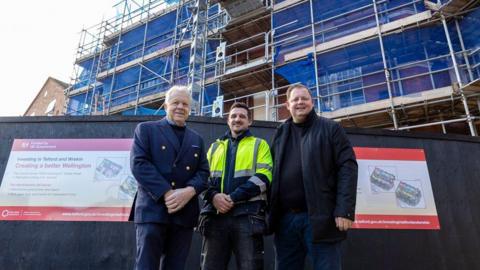 Three men are stood in front of a large building that is covered in metal scaffolding and blue netting. The one on the left has a navy jacket on and short white hair. The man in the middle has brown hair and a yellow, black and grey hi-vis jacket on. The last on the right has a black coat and black top on, and has short light brown hair.