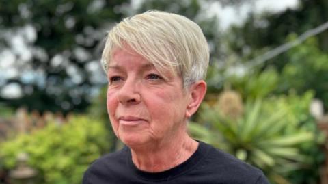 A woman looks to the left of the camera, she has short blonde hair and wears a black T-Shirt. Behind her are some bushes and trees.