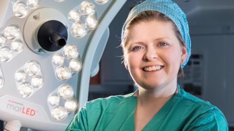 Dr Andrea Woolner wearing surgical scrubs and hair covering, standing in operating theatre looking into the camera