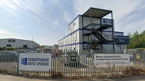 A grey and blue building is seen in the background with a stairwell in front of it. In front of the building is a fence with many posters including one saying Transpennine Route Upgrade, thieves beware and we are registered with the Considerate Constructors Scheme.