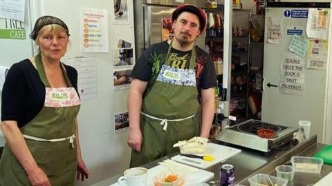 Two volunteers in the kitchen looking at the camera wearing cooking overalls