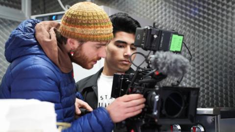 Two men are looking intently into the view finder of a camera. The man on the left, who has a beard, is wearing a blue jacket and a woolen hat. The other man is one of the students. He has dark hair, a black jacket and white T shirt.