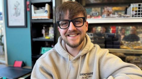 Aaron Stewart-Gillespie has short brown hair with a fringe and wears thick black-rimmed glasses. He has a short beard and is smiling, sitting in a cafe, wearing a cream hoodie.