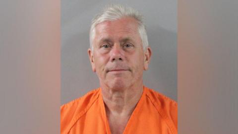 A man in his sixties with white hair looks at the camera during his police mug shot.