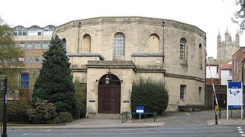 The exterior of Gloucester Crown Court, a round building made of stone with large brown wooden doors outside. By the doors, there are trees and shrubbery. Gloucester Cathedral can be seen in the distance.