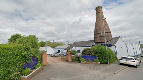 The entrance to a factory site off the side of a road. Blue signs outside the entrance, surrounded by hedges, read "Moorcroft". A tall brick chimney perches on top of one of the buildings.