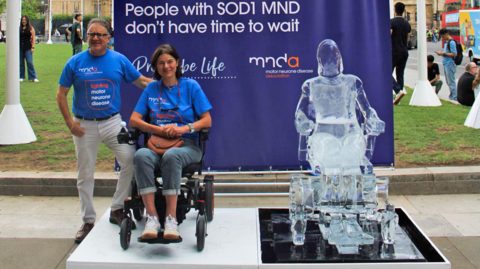A man in a blue tshirt and white trousers standing outdoors next to a woman with dark hair and a blue tshirt in a wheelchair on a platform with a blue background and an ice sculpture of a person in a wheelchair