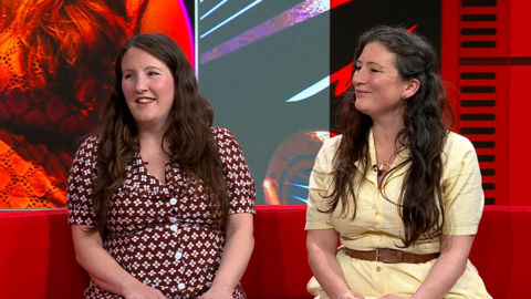 Becky and Rachel Unthank sit next to each other on a red sofa in front of a colourful background. Both have long, dark, wavy hair. They are smiling and looking over to the left of the image. The woman on the left is wearing a maroon dress with a white flower pattern, and the woman on the right is wearing a pale yellow dress with a brown belt.