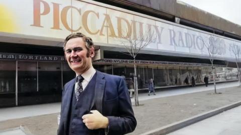 Philip Birch stands outside Piccadilly Radio's studios. He's wearing a suit including waistcoat. The Piccadilly Radio 261 text and logo adorns the outside window behind him.