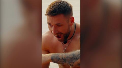 A topless man looks stunned. He has tattoos and a necklace.