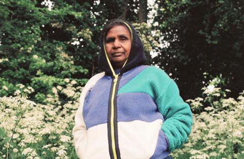 Anupama Gamanagari's mother stands in the Staffordshire countryside. She is wearing a purple, white and green fleece with the black hood up. She is also wearing jeans and stands amid greenery in front of trees.