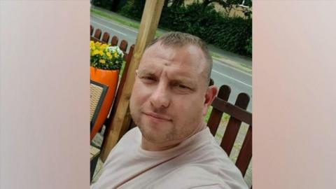 A man with short brown hair who is muscular has a pale pink top on and is looking into the camera with a serious expression. Behind him is a wooden fence and then a road behind that. He appears to be sat down at a table.