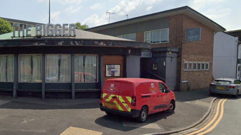 A brick built two storey building with a curved glazed front. Large letters on the roof of the curved frontage spell out The Rigger.