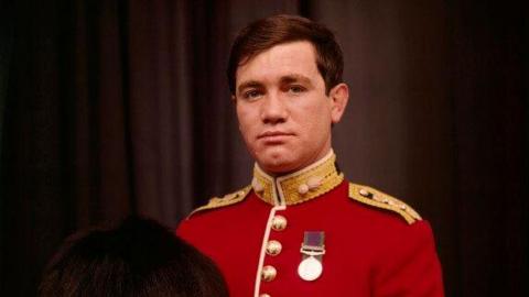 A cropped version of a National Portrait Gallery image of the Army officer Captain Robert Nairac.  He has short, brown hair and is wearing a red military jacket decorated with gold buttons, gold epaulettes and single medal pinned to his chest.  He is holding a tall, black bearskin hat.