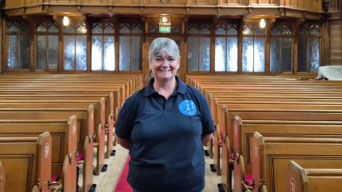 Maureen Leonard standing in a church's sanctuary, in the middle of the pews. She has short, grey hair and is wearing a blue T-shirt
