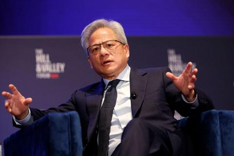 Jensen Huang, boss of Nvidia, sits in a chair with his hands out as he explains something to an audience. He is wear a blue suit, with a shirt and dark tie.
