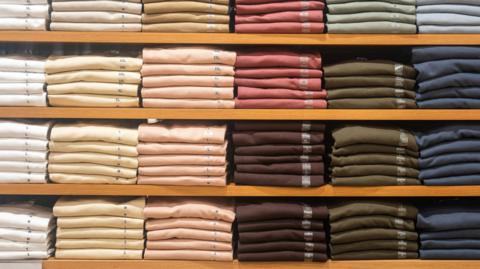 Piles of folded clothing in white, yellow, peach, pink, burgundy, green and blue are folded on the shelf in clothing store. 