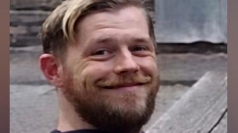 A man in his late 30s, with blonde hair combed to the side and a ligh brown beard, smiles at the camera. He is wearing a blue hooded jumper. 