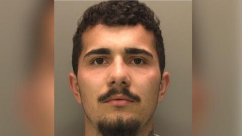 A man with short dark hair, slightly curly, with a black moustache and goatee beard is looking straight into the camera of a police custody picture.