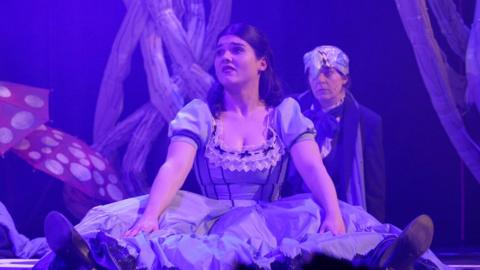 A girl sitting on a stage floor, wearing a blue Alice in Wonderland dress and looking confused. 