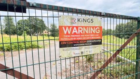 A section of land which is guarded by a tall metal gate. On the gate is a sign for the security company, which reads 'Warning - these premises are monitored and patrolled by Kings Security personnel'.